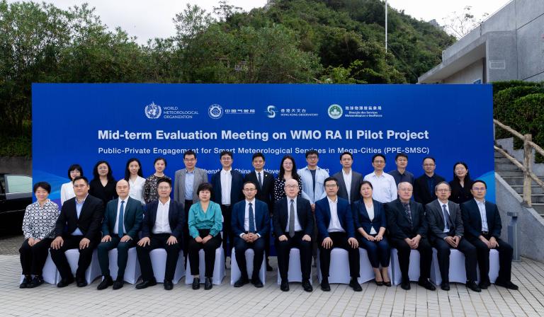 Group of people posing for a photo at the mid-term evaluation meeting on wmo ra ii pilot project event, standing in front of a banner.